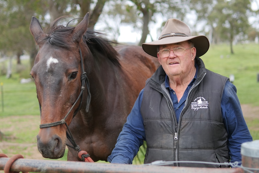 A man wearing a hat stands next to a horse, they are outside on a property with trees.