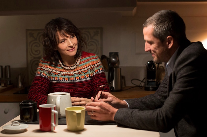 A brunette woman in bright patterned jumper sits at kitchen table at night slightly smiling at seated man.