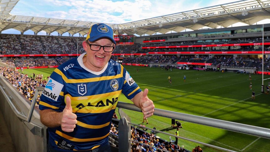 A Parramatta Eels fan gives two thumbs up from the stands of a football stadium.