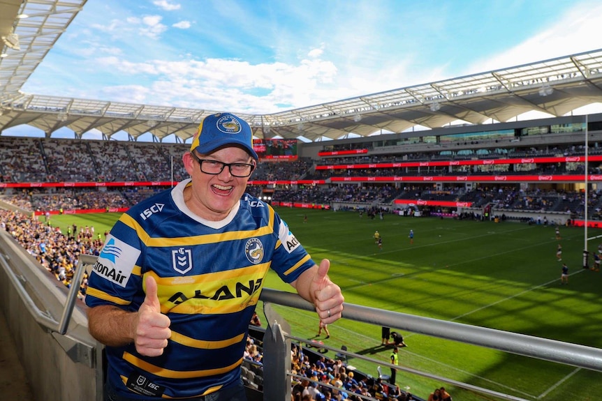 A Parramatta Eels fan gives two thumbs up from the stands of a football stadium.