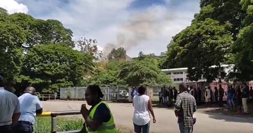 Small crowd watches on as black smoke billows from a burning hut that is just visible from behind trees.