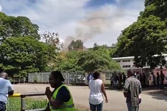 Small crowd watches on as black smoke billows from a burning hut that is just visible from behind trees.