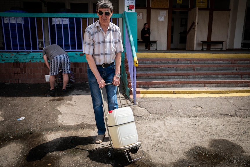 A man wearing jeans, a checked shirt and sunglasses pulls a short metal trolley with a plastic water container strapped to it 