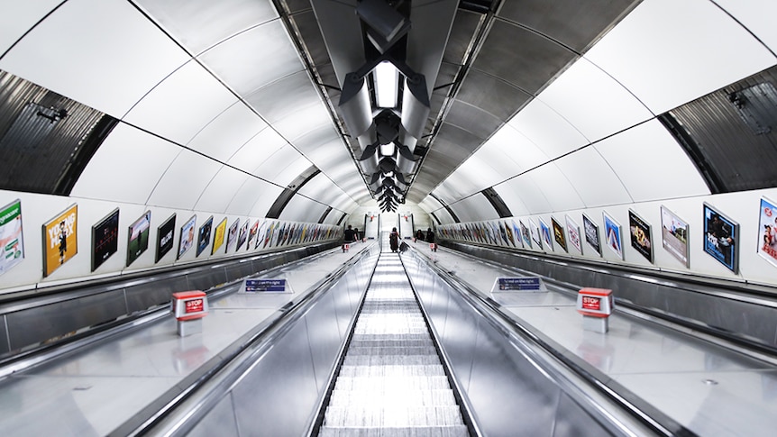 A steep London Tube escalator looking from the top down with the walls on either side lined with colourful posters