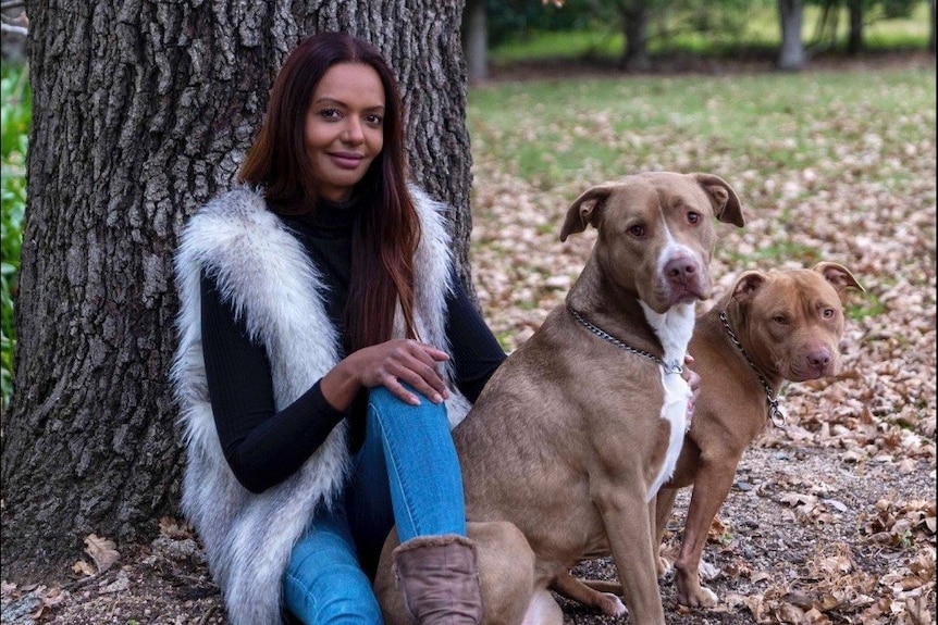 A woman with dark hair sits under a tree with two dogs