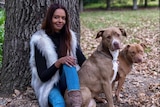A woman with dark hair sits under a tree with two dogs