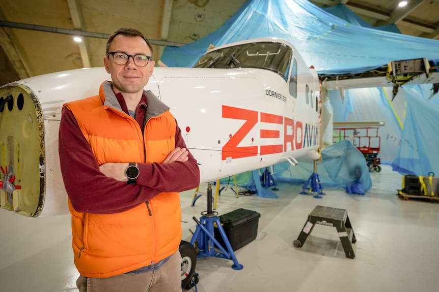 A man wearing a fluro orange vest crosses his arms in front of a plane.