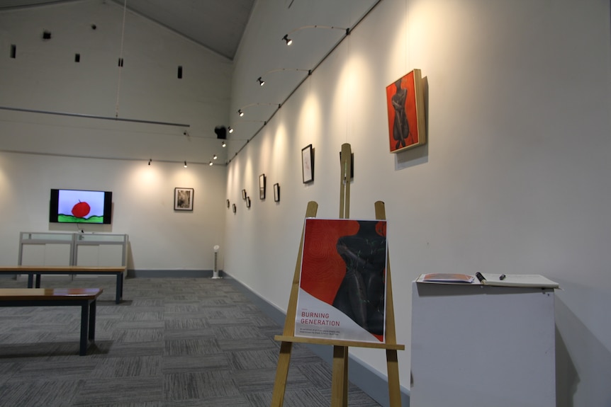 A gallery with numerous artworks on the wall, and a poster on an easel with the words 'Burning Generation' 