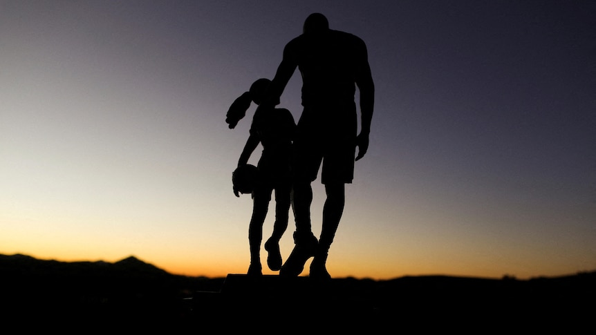 A statue of a man with his arm around a young girl holding a basketball with the sun setting over mountains.