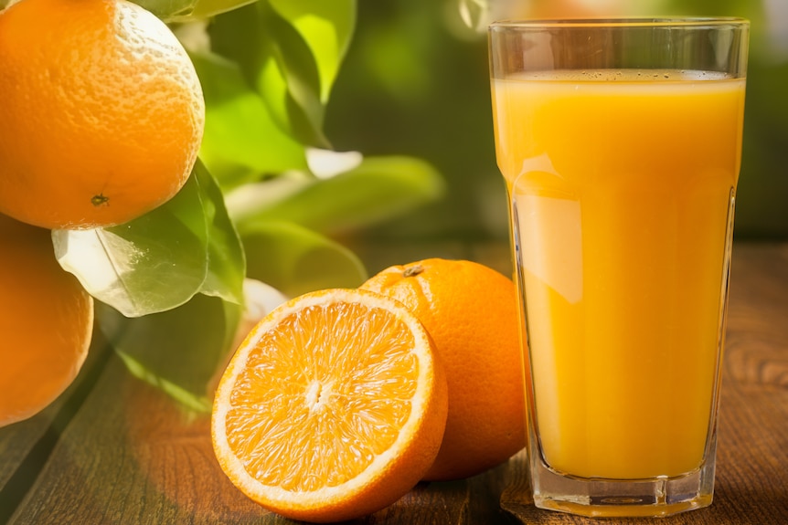 Oranges, including one sliced in half, sit next to a tall glass of orange juice.