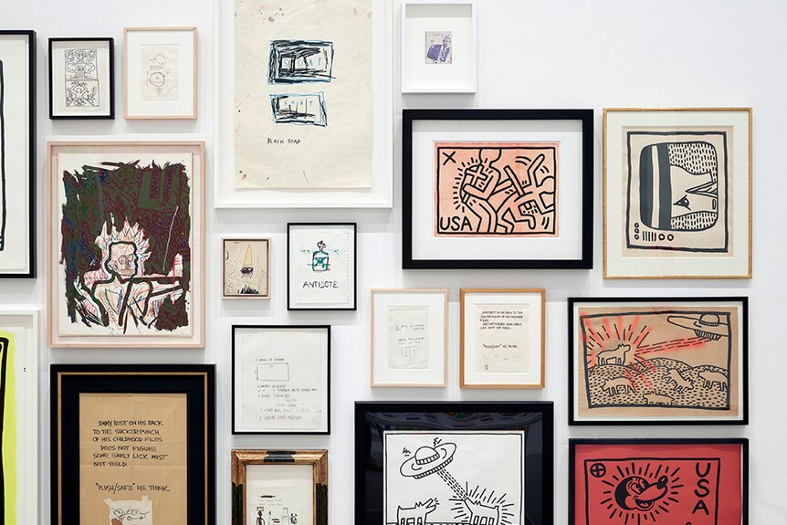 Various Keith Haring and Jean-Michel Basquiat artworks in frames displayed tightly together on white gallery wall.