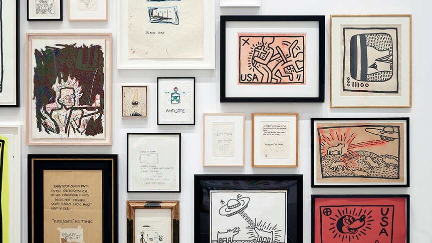 Various Keith Haring and Jean-Michel Basquiat artworks in frames displayed tightly together on white gallery wall.