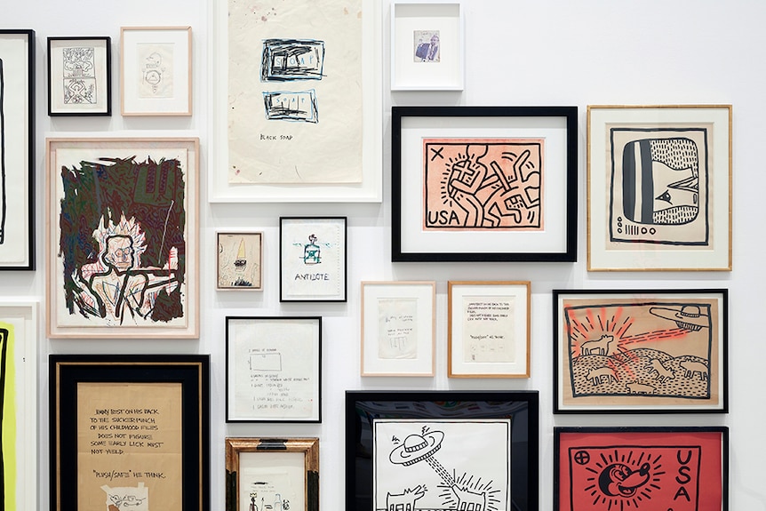 Various Keith Haring and Jean-Michel Basquiat artworks in frames displayed tightly together on white gallery wall.