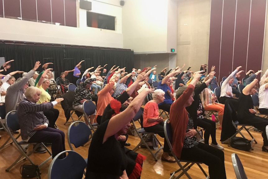 About 100 seniors sitting on chairs, with their arms stretched overhead and leaning to the side.