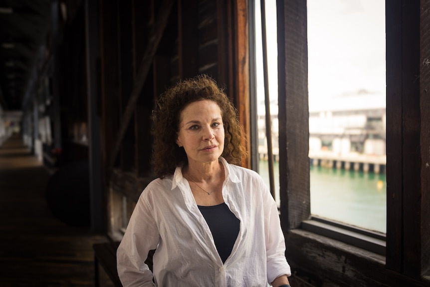 Sigrid Thornton, a 64-year-old white woman with curly brown hair wearing a white blouse and black pants stands beside wood panel