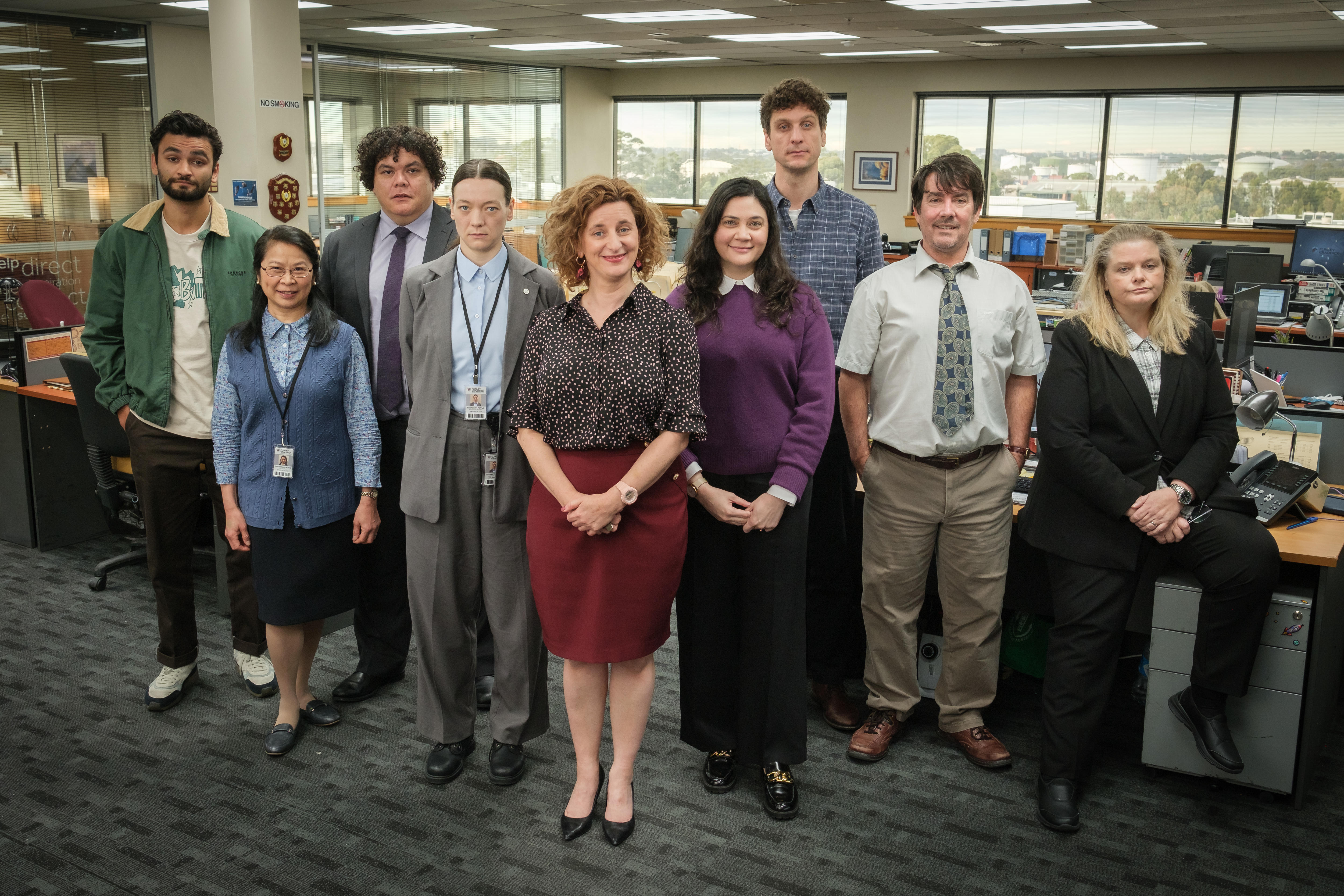 A group of nine people, Felicity Ward centre, stand in an open-floor office with a window behind them and pose for the camera.
