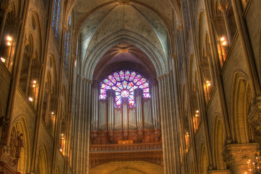 A colourful stained glass window and towering organ pipes sits near the roof of a large cathedral