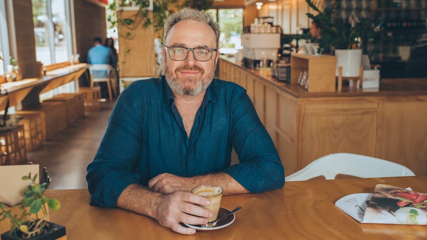 A man with curly grey hair and a beard, wearing a blue shirt and glasses, sitting at a table in a cafe.
