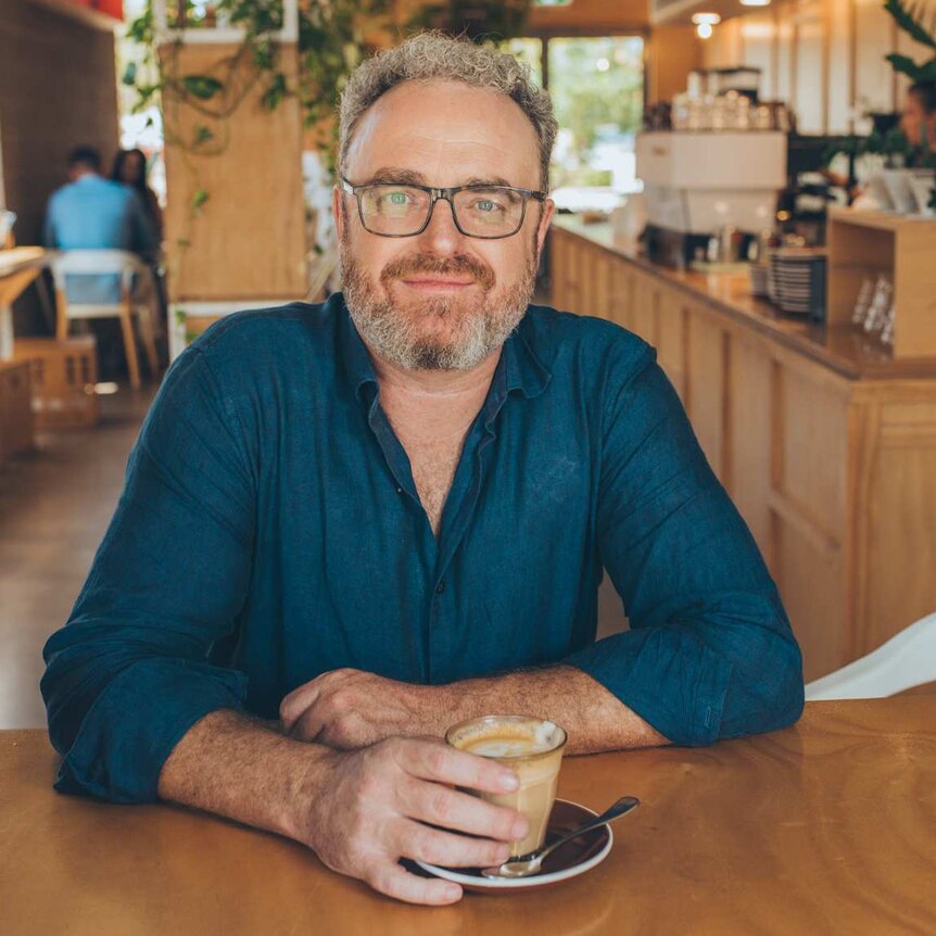 A man with curly grey hair and a beard, wearing a blue shirt and glasses, sitting at a table in a cafe.