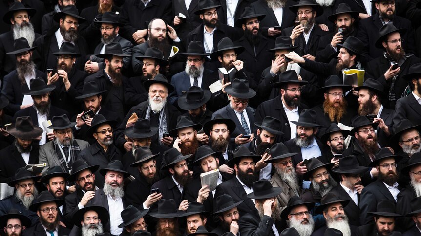 Dozens of bearded men wearing black coats and hats gather for a group photo.