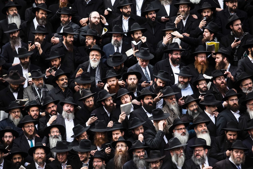 Dozens of bearded men wearing black coats and hats gather for a group photo.