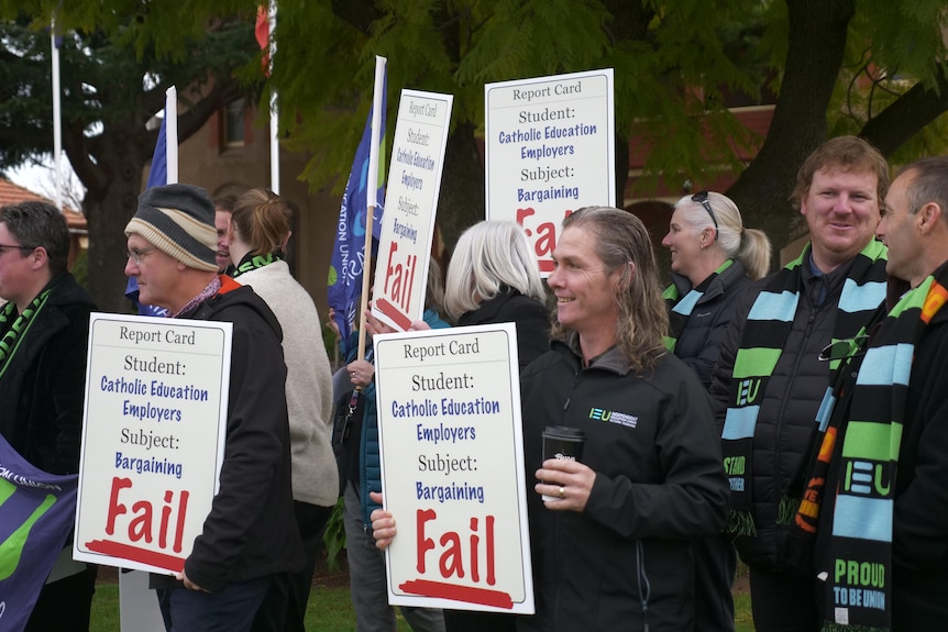 Protestors holding placards and wearing union-branded scarves stand on a school lawn. 