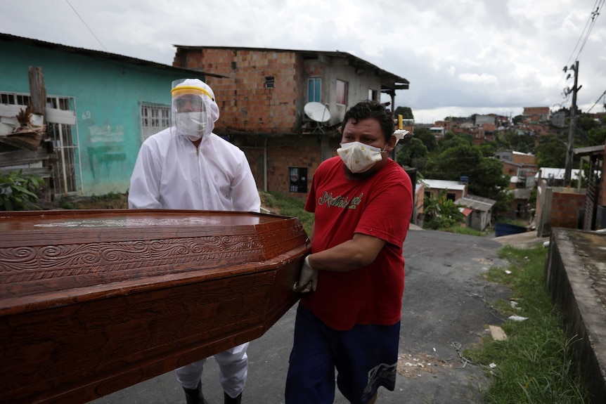 Two men in protective equipment carry a coffin along a street, with a favela in the background.