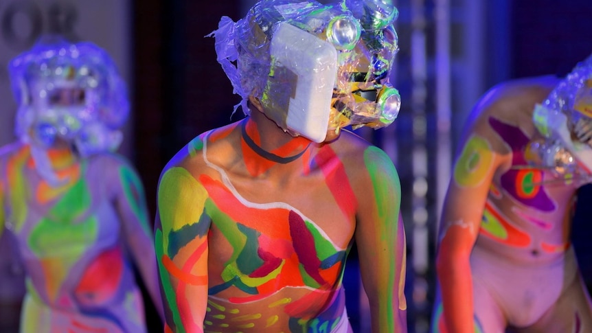Stage shot, with head to waist torso of dancer in foreground, wearing mask made from trash and bare torso painted colourfully.