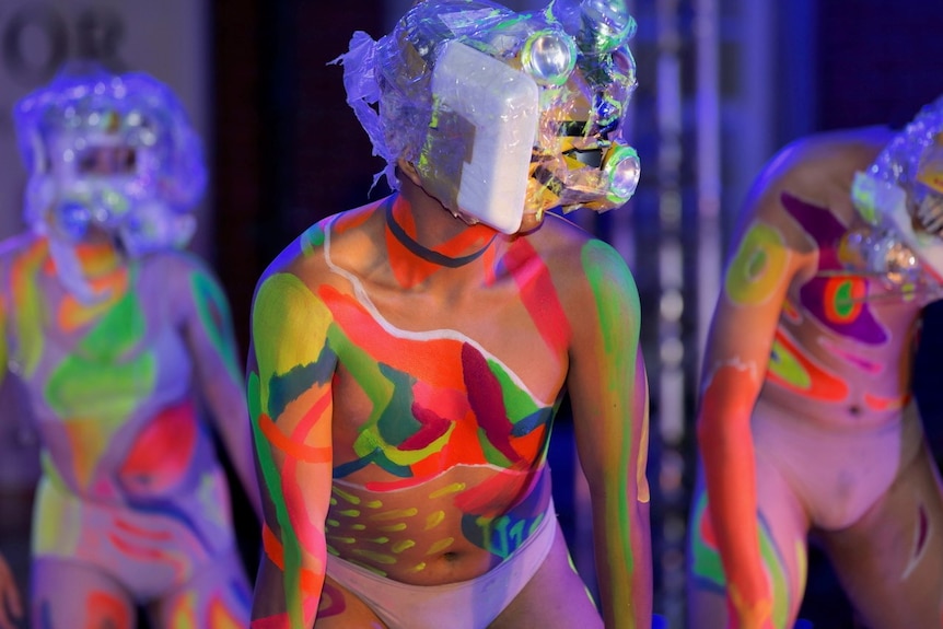 Stage shot, with head to waist torso of dancer in foreground, wearing mask made from trash and bare torso painted colourfully.