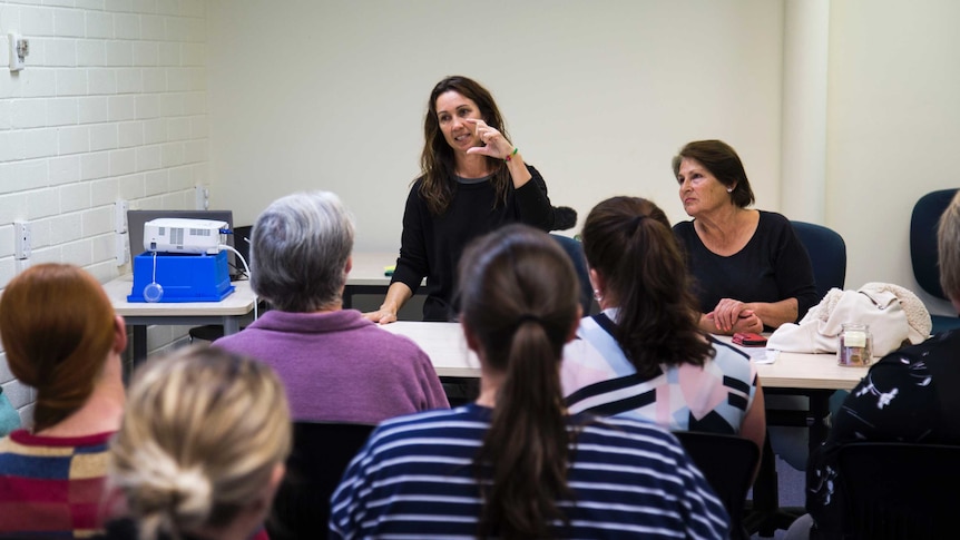 A woman stands at the front of a group of seated people, showing them sign language