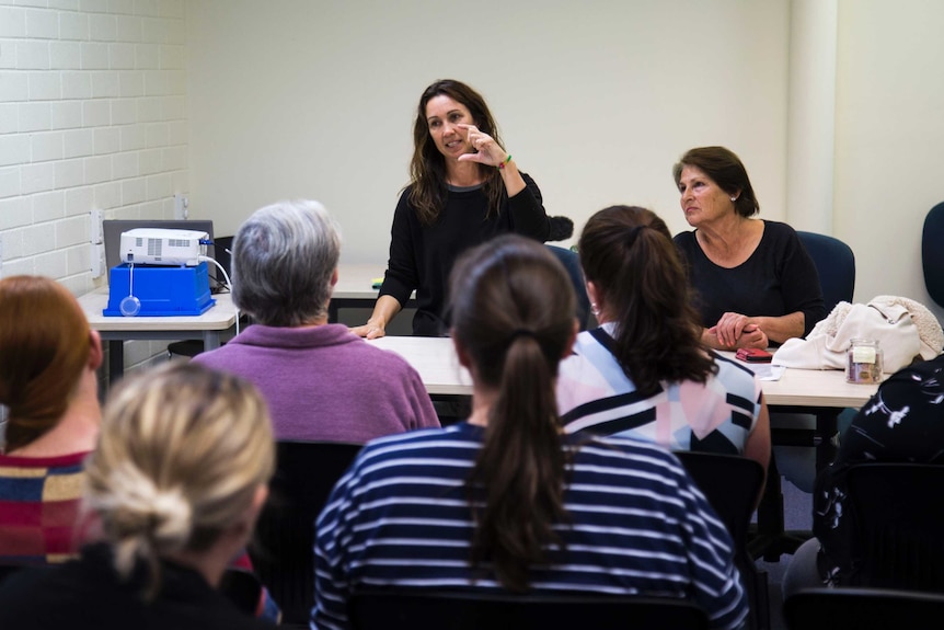 A woman stands at the front of a group of seated people, showing them sign language