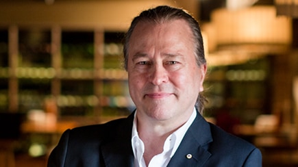 Neil Perry in a dark suit jacket leaning on a table in a restaurant interior.