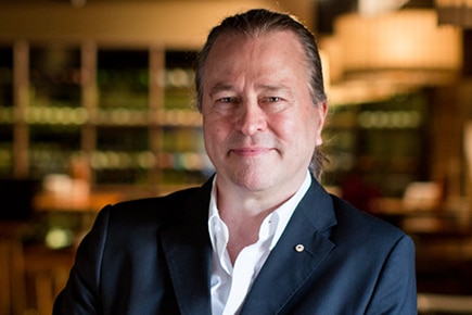 Neil Perry in a dark suit jacket leaning on a table in a restaurant interior.