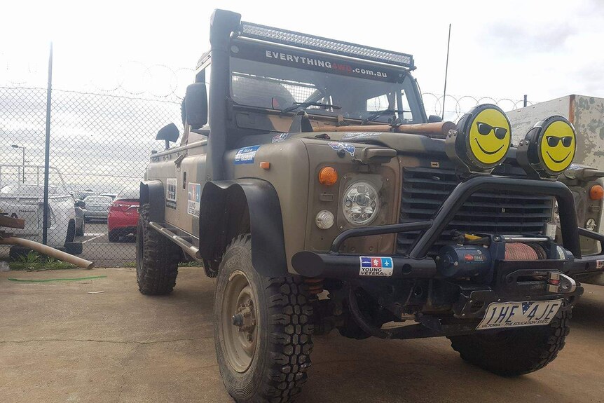 A Landrover painted in camouflage and covered in stickers, with smiley faces on the headlights sits in a compound