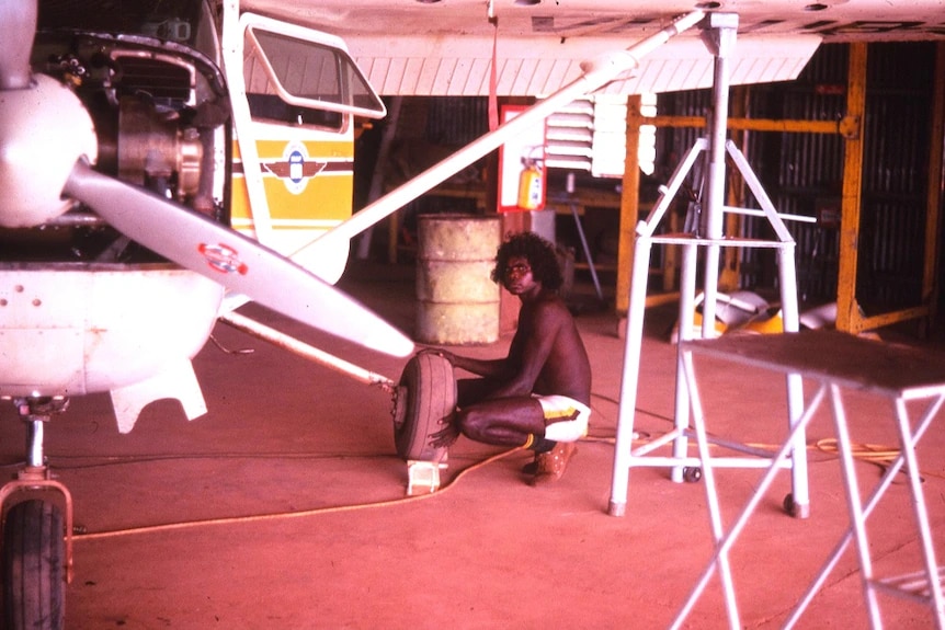 Man squats beside small aeroplane holding tyre.