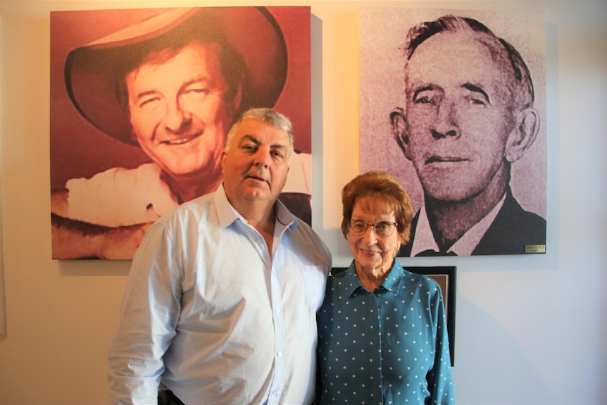 Elderly woman, green dress, glasses, man , grey hair, shirt, in front of large pics, one man wears akubra, other b & w in suit.