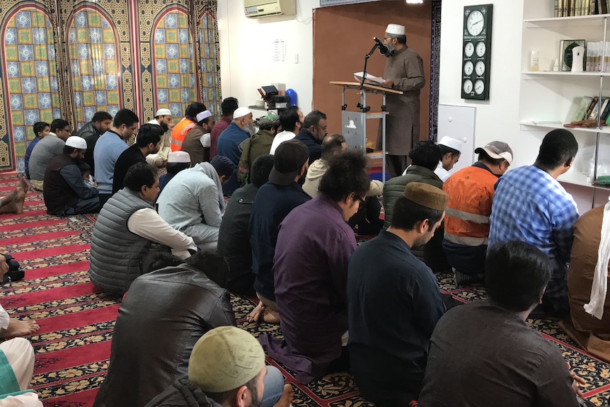 A group of men sitting down in the mosque prayer room with the Imam standing while speaking