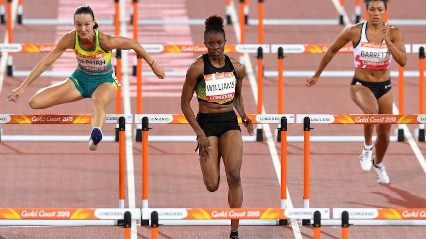 Three female athletes shown mid-race with the athlete on the left in the air while jumping over a hurdle