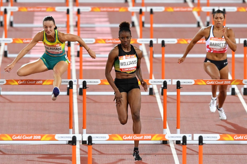 Three female athletes shown mid-race with the athlete on the left in the air while jumping over a hurdle