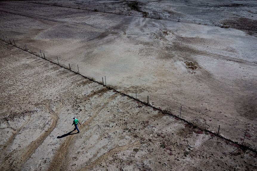 A drone shot of Boyd Webb walking near a tall fence, with bare arid land stretching into the distance.
