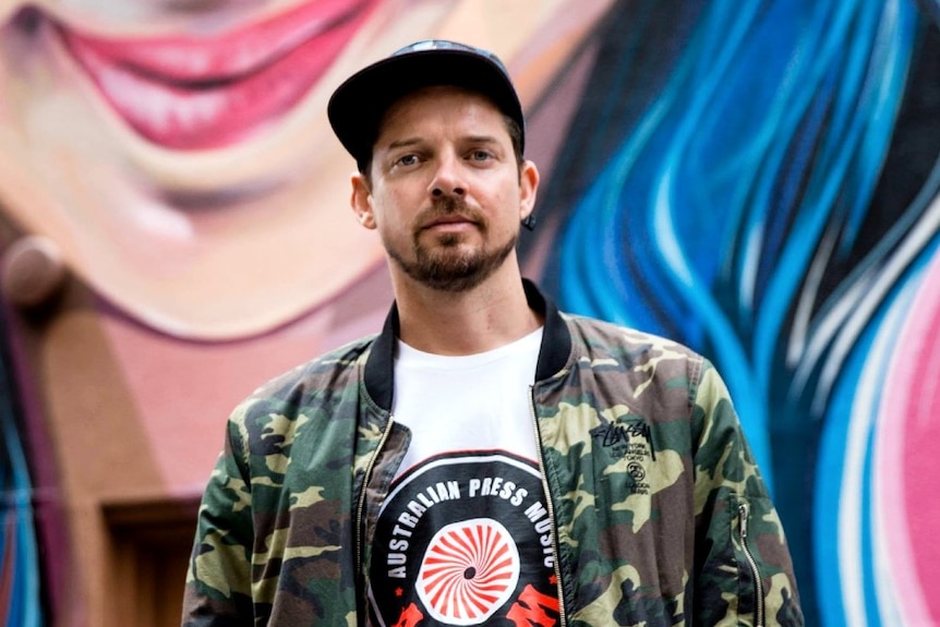 A man in a camouflage jacket stands in front of one of his colourful mural portraits