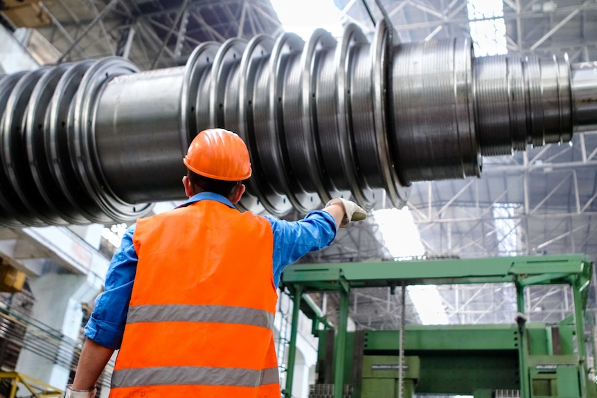 A male worker in a high-vis vest and hard hat points at machinery