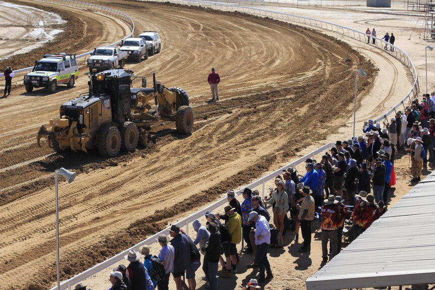 A big yellow machine drags dirt on a racetrack as a crowd watches.
