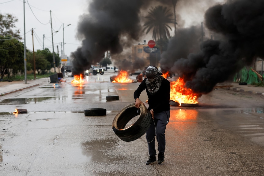 A man carrying a tyre with flames in the background