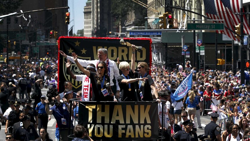 US women's footballers wave to a huge crowd from the back of a bus, with a sign on it saying 'Thank you fans'.