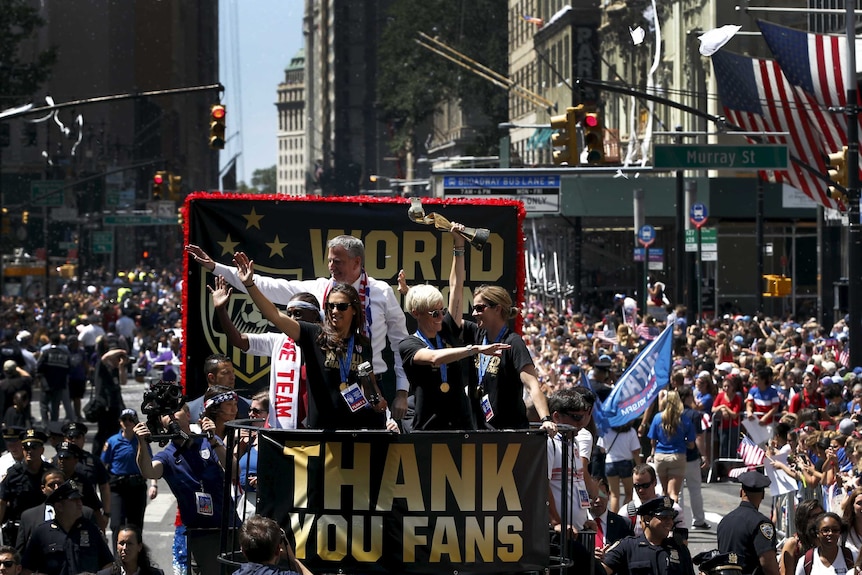 US women's footballers wave to a huge crowd from the back of a bus, with a sign on it saying 'Thank you fans'.