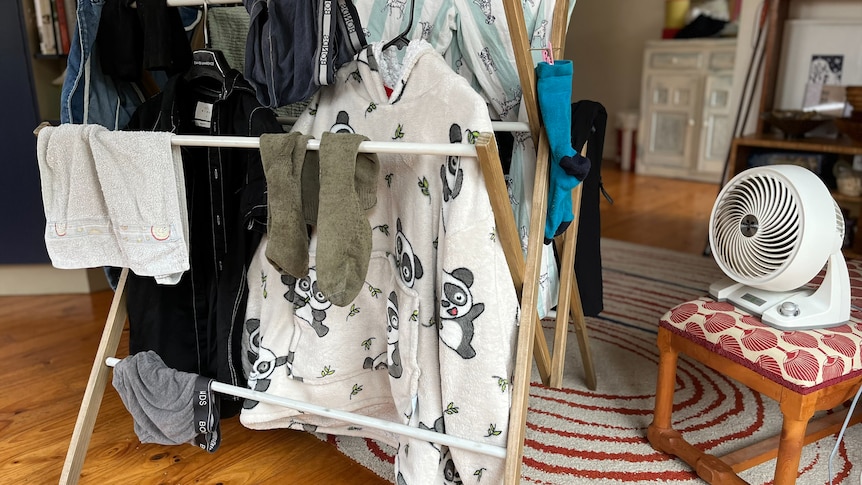 Clothing hangs on a dryer rack indoors with a small fan nearby