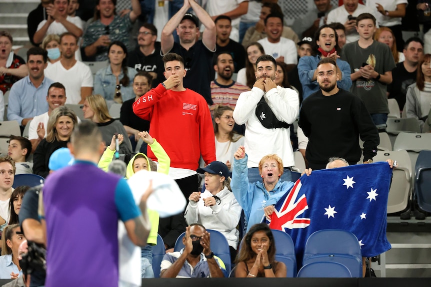 Tennis player Nick Kyrgios is blurry in the foreground on court and cheered on by fans in the background.