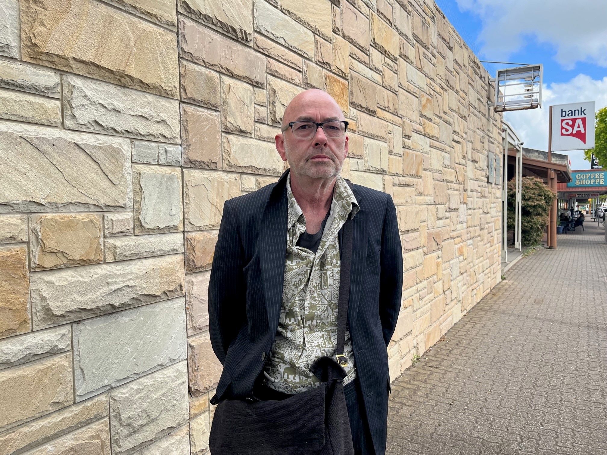 A bald man wearing a suit and floral shirt standing outside a sandstone wall