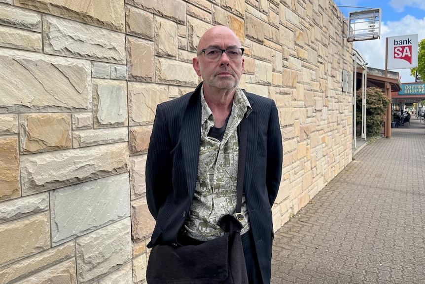 A bald man wearing a suit and floral shirt standing outside a sandstone wall